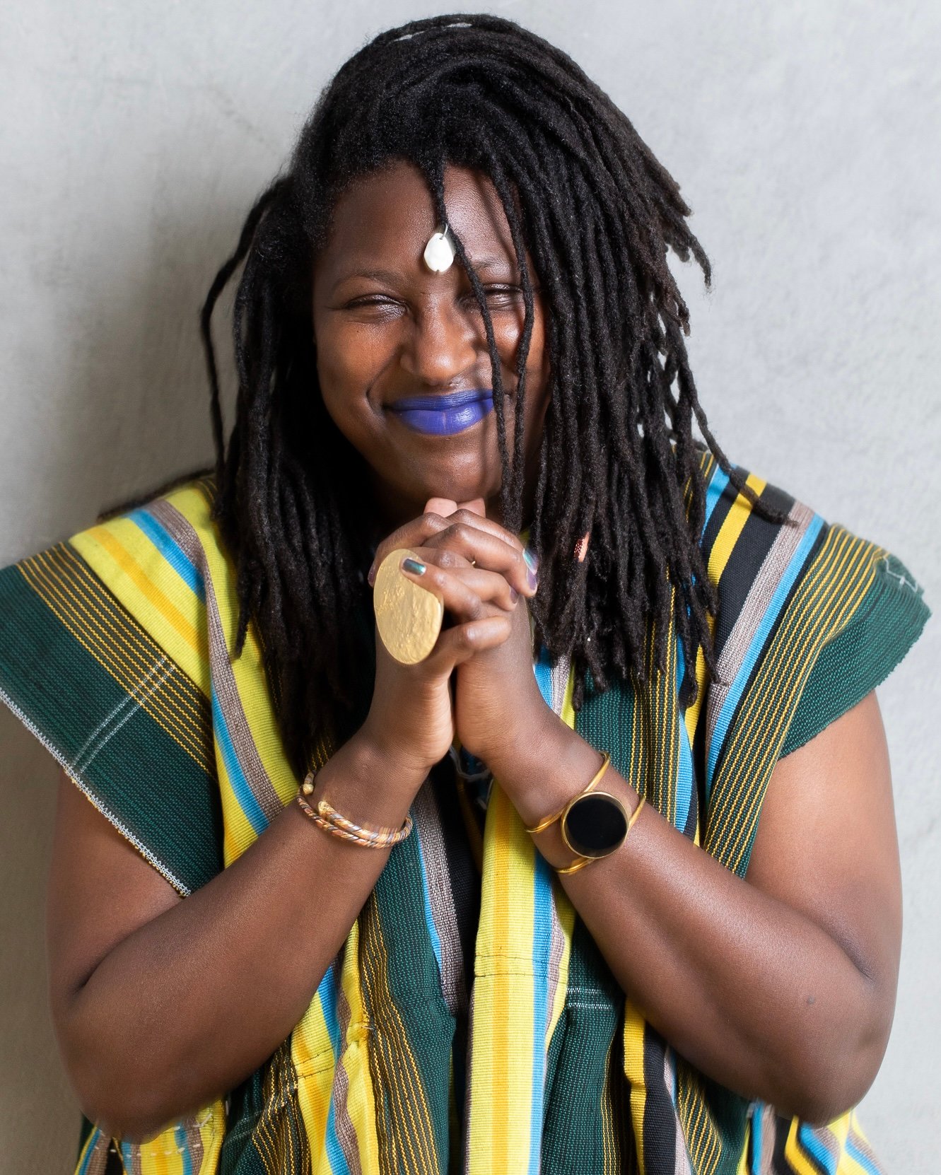 Portrait de Mame Marieme Lô femme noire, souriant, avec du rouge à levres bleu et une veste sans manches à rayures bleues vertes et jaunes elle a des locks mi-longues